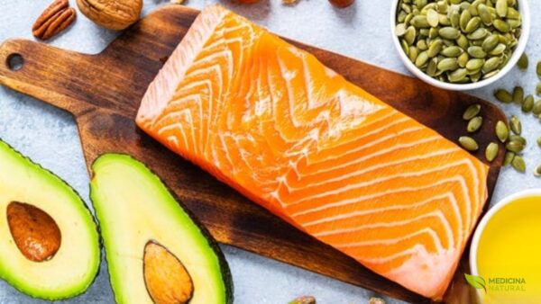 Composição harmoniosa de alimentos ricos em gorduras boas: salmão fresco, abacate cortado ao meio, variedade de oleaginosas (nozes, amêndoas, pistache), sementes de abóbora em tigela de madeira e azeite de oliva em pequeno recipiente. Dispostos sobre superfície clara, representando as principais fontes de ácidos graxos insaturados essenciais para saúde cardiovascular e cerebral.