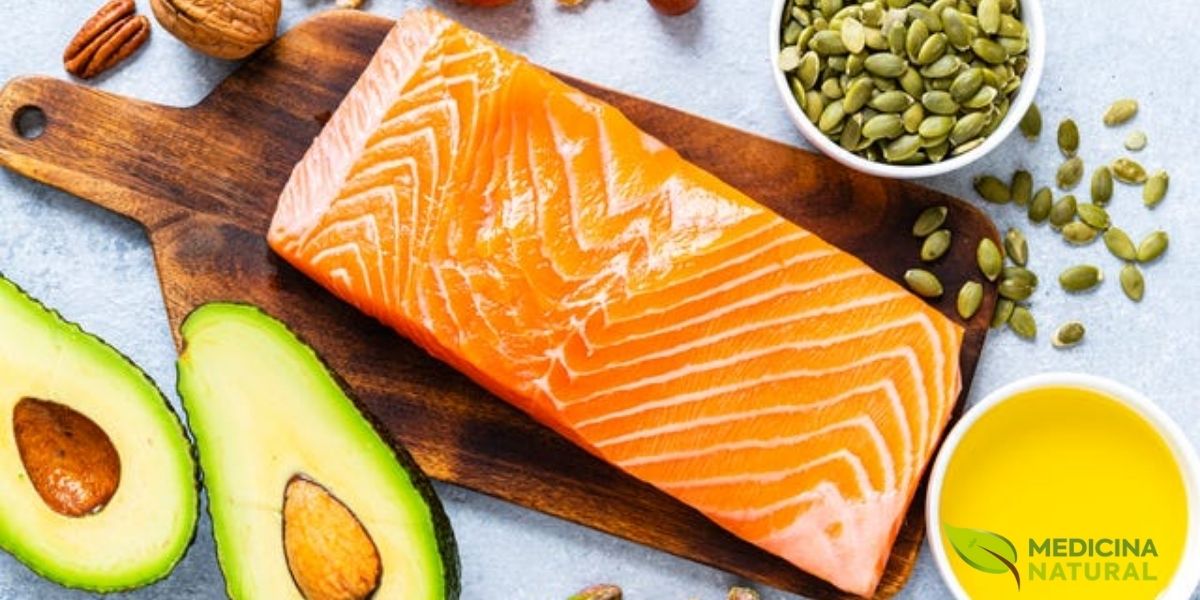 Composição harmoniosa de alimentos ricos em gorduras boas: salmão fresco, abacate cortado ao meio, variedade de oleaginosas (nozes, amêndoas, pistache), sementes de abóbora em tigela de madeira e azeite de oliva em pequeno recipiente. Dispostos sobre superfície clara, representando as principais fontes de ácidos graxos insaturados essenciais para saúde cardiovascular e cerebral.