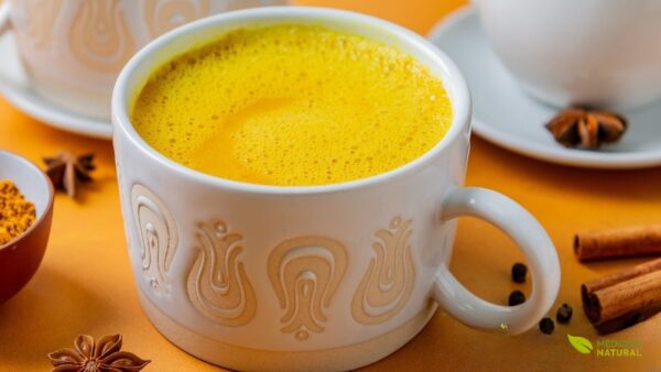 Xícara de vidro com chá de cúrcuma Golden Milk decorado com canela em pó, acompanhado de paus de canela sobre superfície clara. Esta versão aromática combina as propriedades anti-inflamatórias da cúrcuma com o calor da canela, criando uma bebida reconfortante e terapêutica. Ideal para fortalecer o sistema imunológico e melhorar a digestão.