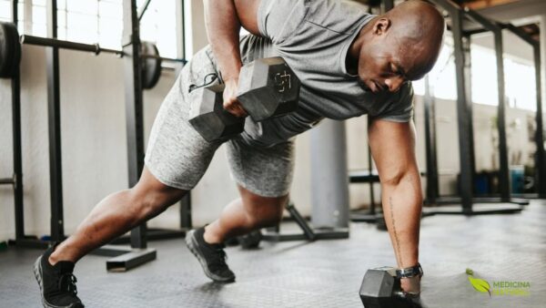 Atleta executando exercício composto com halteres em academia moderna, demonstrando a versatilidade dos treinos com pesos livres para o desenvolvimento muscular completo. Os exercícios com halteres permitem uma maior amplitude de movimento e recrutamento de fibras musculares estabilizadoras, gerando um estímulo mais completo para a hipertrofia. A nutrição pós-treino, incluindo proteínas de rápida absorção e carboidratos, é crucial para otimizar a recuperação e maximizar os ganhos de massa muscular. Fonte: Muscle & Fitness.